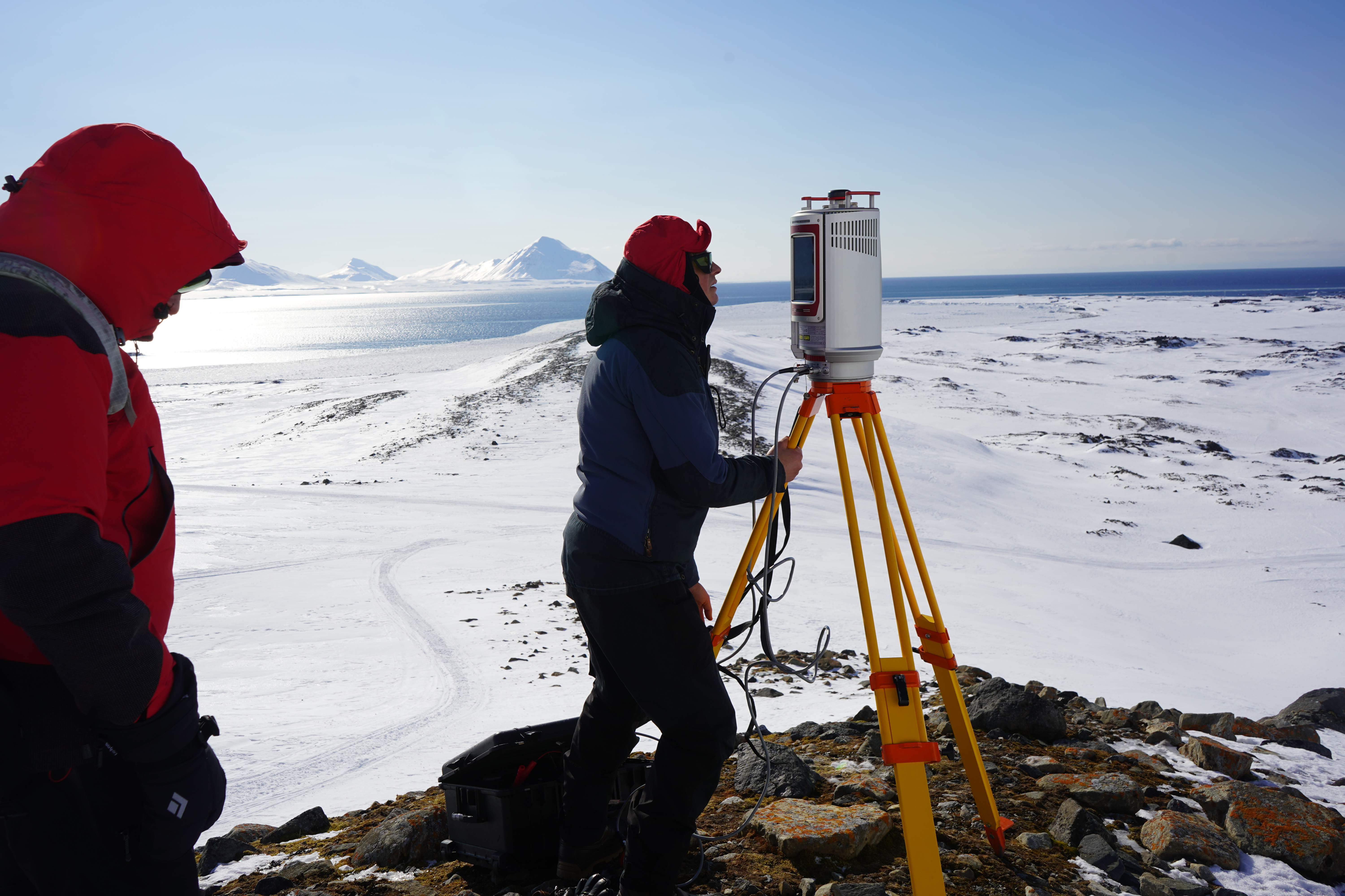 A person standing next to a scientific instrument with snow, sea and mountains in the background