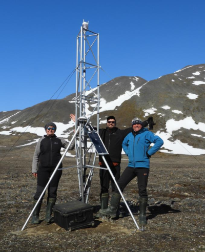 "Photograph of three people standing next to an instrument tower on tundra with mountains in the background"